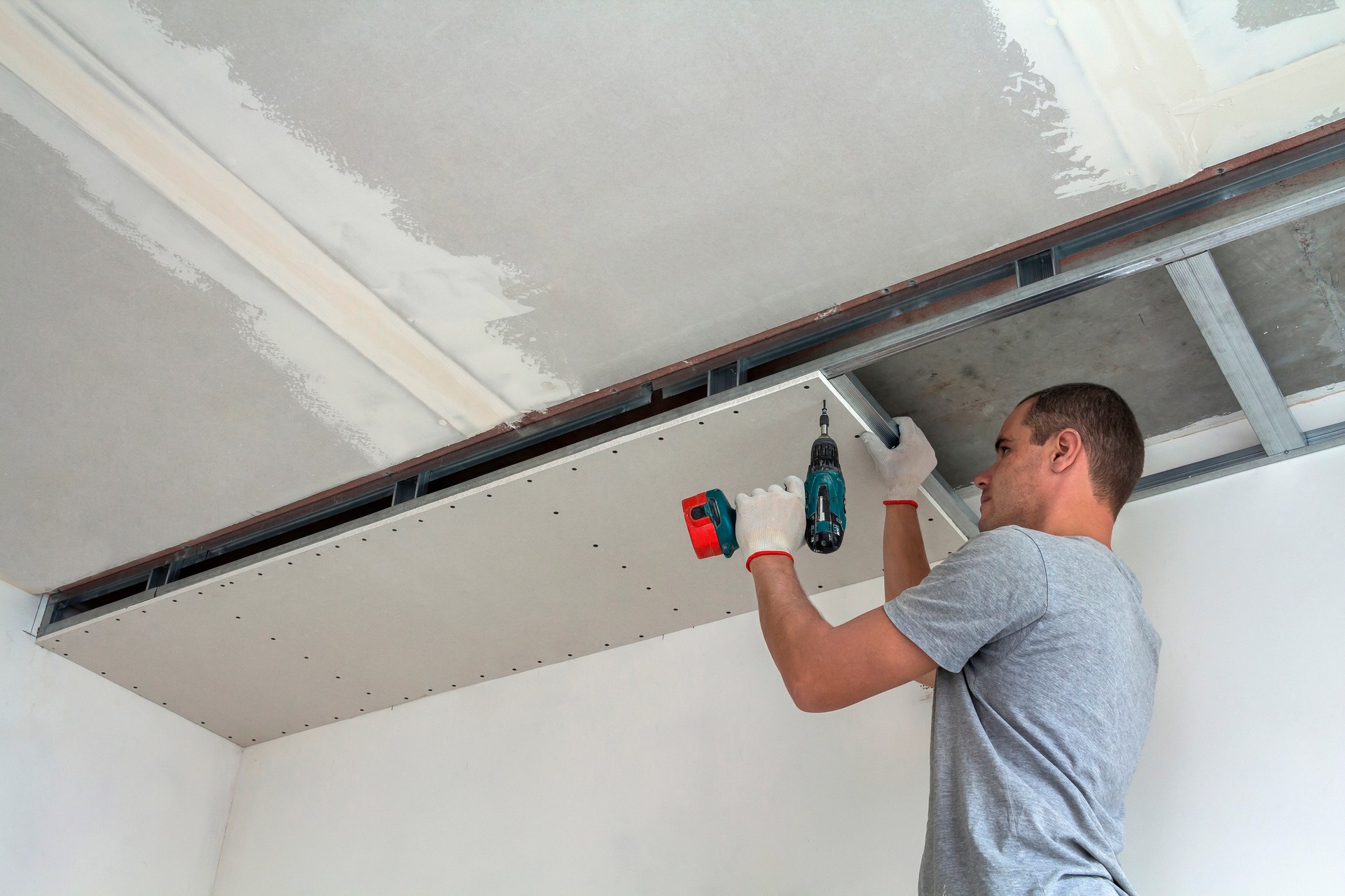 Hombre joven en ropa habitual y guantes de trabajo de fijación de paneles de yeso suspendido techo a marco de metal utilizando un destornillador eléctrico en el techo aislado con papel de aluminio brillante. DIY, hágalo usted mismo concepto.